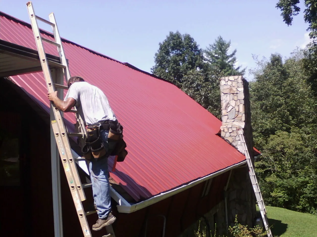 Roofer on a ladder beside a red metal roof and stone chimney — Soffit & Fascia Repair by Roofing Brothers in Lorraine